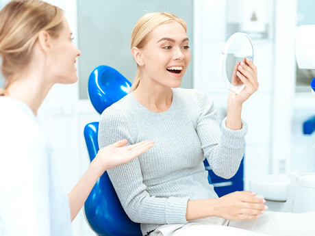 woman with blonde hair in grey sweater smiling at her reflection next to dentist